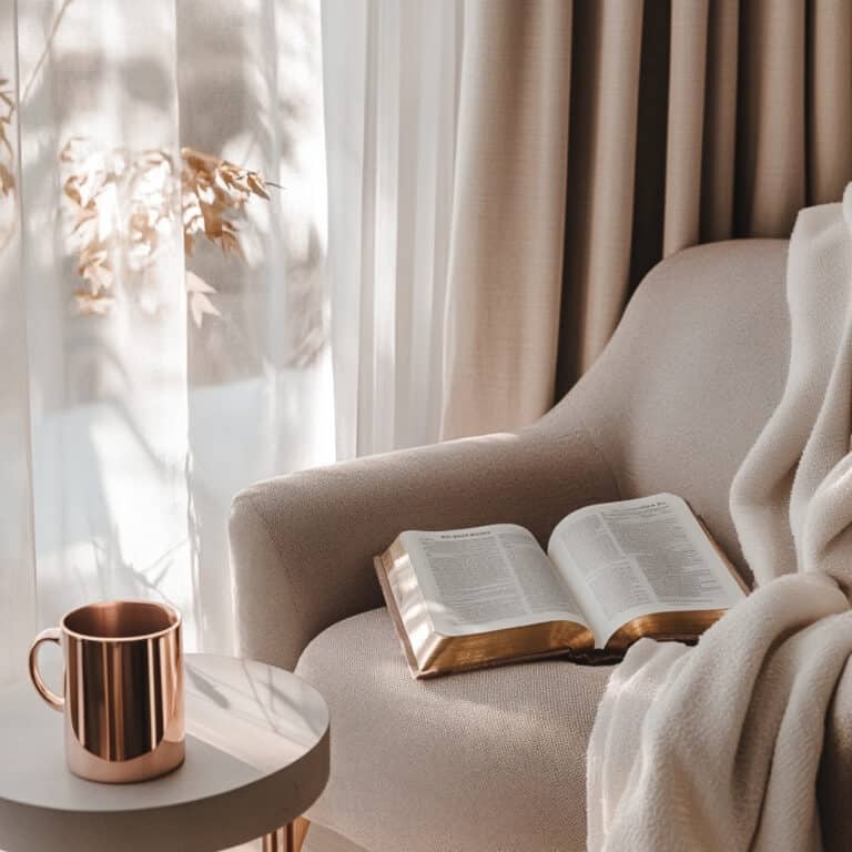 Open Bible on a soft beige armchair beside a rose gold coffee mug on a gray table, with cozy blanket and sheer curtains letting in warm morning light. Minimalist, faith-inspired reading nook with calming gray and rose gold accents.