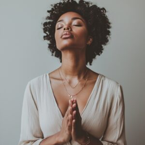 Modern woman in a cream blouse with eyes closed and hands pressed in prayer, expressing bold faith and spiritual stillness in a minimalist setting